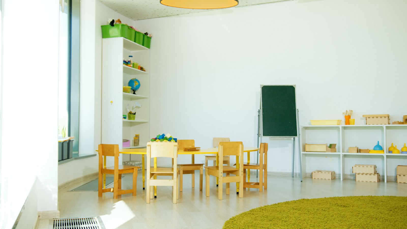 A bright, tidy classroom with small wooden tables and chairs, open shelves of toys and a green rug