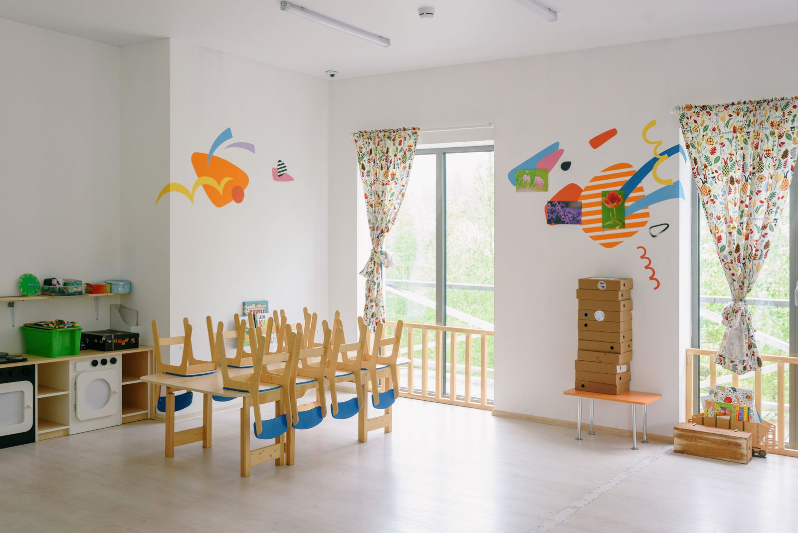 A sunny classroom with low wooden chairs set at a table, a play kitchen and curtains at tall windows
