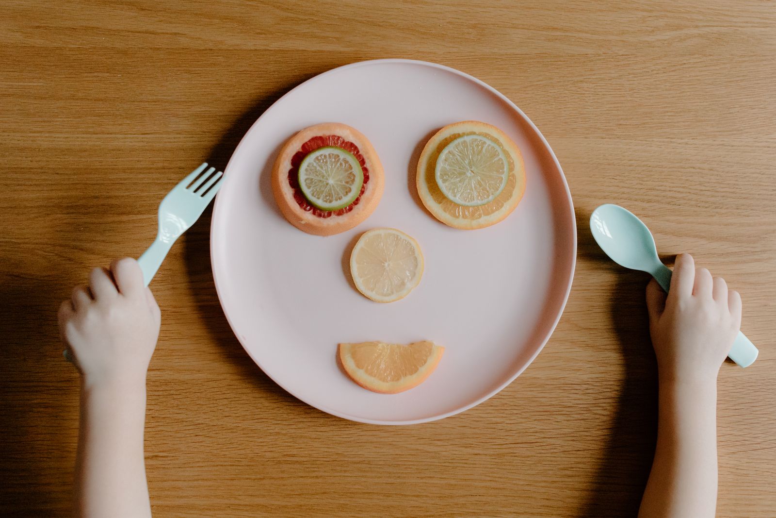 A plate with orange and lemon slices arranged like a face, with a child’s hands holding a fork and spoon